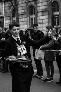 Photographie en noir et blanc d'un serveur saluant la foule durant la Course des Cafés à Paris, reportage de rue et interaction avec le public.