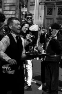 Photographie en noir et blanc de la Course des Cafés à Paris, un serveur en gilet passant devant des spectateurs prenant des photos, reportage urbain authentique.