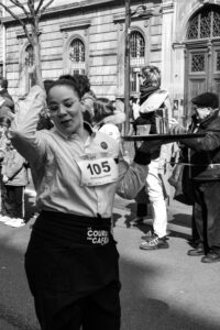 Suggestion : Photographie en noir et blanc d'une serveuse concentrée lors de la Course des Cafés à Paris, portrait de rue authentique et reportage événementiel.