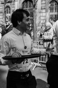 Suggestion : Portrait en noir et blanc d'un serveur souriant avec son plateau lors de la Course des Cafés à Paris, reportage photo lifestyle et traditions parisiennes.