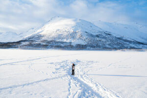 Homme marchant sur un lac gelé en Norvège entouré de montagnes enneigées sous un ciel bleu.