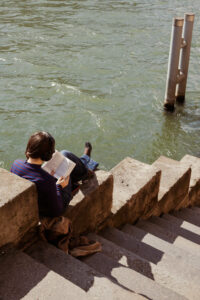 Vue de haut d'une personne assise sur les marches en pierre des quais de Seine à Paris, lisant un livre avec un casque audio face à l'eau.