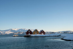 Trois maisons colorées sur un rivage enneigé à Sommarøy, Norvège, avec des montagnes enneigées et un ciel bleu clair en arrière-plan.