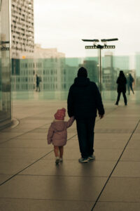 Silhouette de dos d'un père tenant la main de sa petite fille marchant sur une esplanade urbaine moderne et épurée.