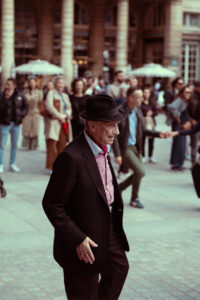 Portrait de profil d'un vieil homme élégant en costume sombre et chapeau noir, marchant sur une place animée à Paris.