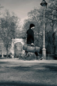 Photographie de rue en noir et blanc d'un homme portant une sacoche et marchant devant un réverbère ancien et une arche dans un parc à Paris.