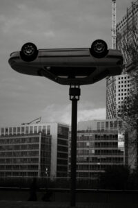 Photographie en noir et blanc d'une sculpture de voiture à l'envers sur un poteau, avec des immeubles de bureaux en arrière-plan.