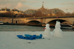 Deux bonhommes de neige et des luges bleues sur les quais de Seine enneigés, avec le Pont des Invalides et le Grand Palais en arrière-plan à Paris.