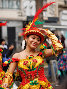 Portrait d'une jeune femme souriante en costume de carnaval rouge et jaune orné de paillettes et de plumes, lors d'un défilé de rue à Paris.