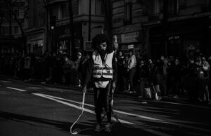 un "Fouettard" de carnaval portant un masque, un gilet de sécurité et tenant des cordes/fouets sur une avenue parisienne.