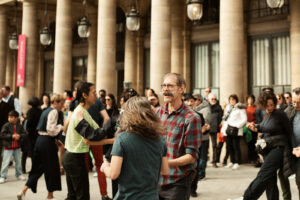 Scène de danse collective en extérieur à Paris, mettant en avant un homme moustachu souriant en chemise à carreaux entouré d'autres danseurs devant des colonnes historiques.
