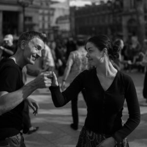 Portrait en noir et blanc d'un homme et d'une femme souriants en train de danser sur une place publique à Paris, se tenant par la main.