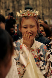 Portrait d'une jeune femme souriante en costume traditionnel hanfu avec une coiffe ornée, tenant des fleurs de cerisier lors d'un défilé à Paris.