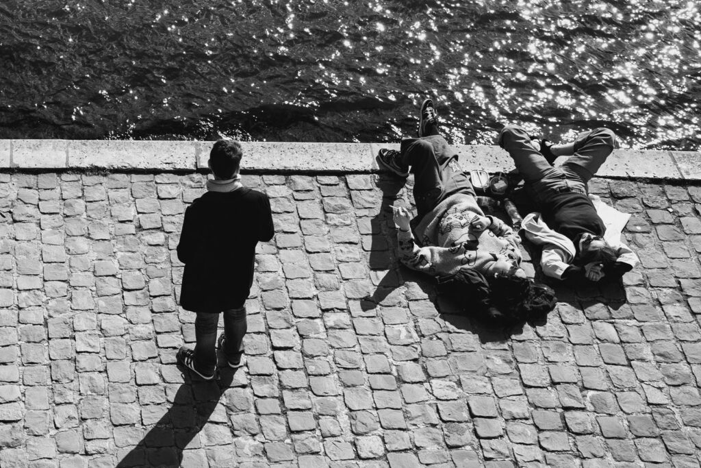 Vue de dessus en noir et blanc de deux personnes allongées sur les pavés des quais de Seine à Paris, à côté d'un passant debout, avec le scintillement de l'eau en arrière-plan.