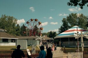 Vue d'une fête foraine d'époque avec une grande roue rouge, un chapiteau rayé bleu et blanc et des panneaux de passage à niveau ferroviaire sous un ciel bleu.