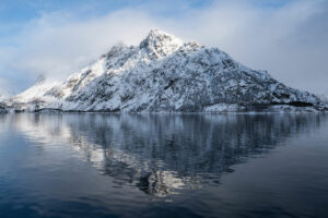Photographie d'une montagne escarpée couverte de neige se reflétant parfaitement dans la surface calme d'un lac ou d'un fjord sous un ciel nuageux.