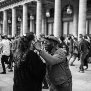 un vieil homme souriant avec une casquette, touchant avec tendresse le visage d'une femme sous des arcades à Paris.